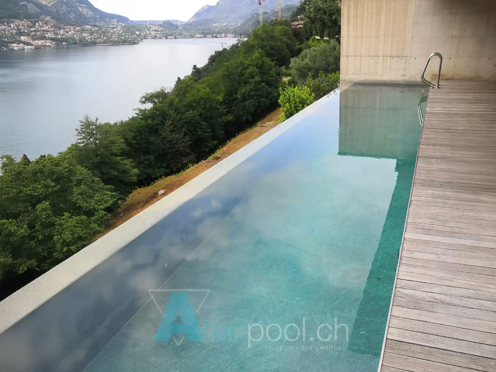 Piscina a sfioro moderna con decking in legno e vista lago – Atec Pool Piscina a sfioro con rivestimento in legno e vista panoramica sul lago
