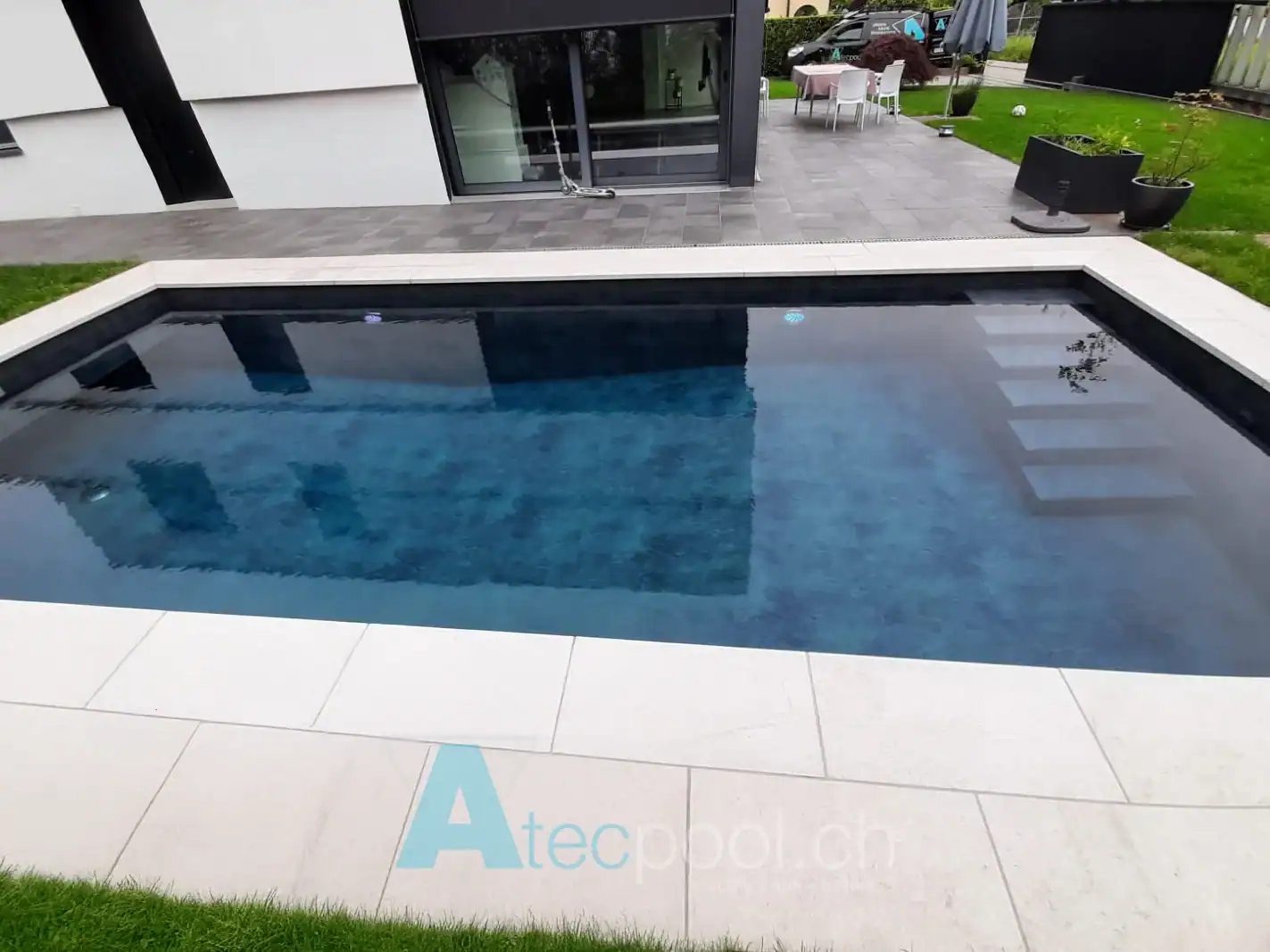 Piscina residenziale moderna con rivestimento scuro e bordo in pietra chiara – Atec Pool Piscina interrata rettangolare con rivestimento scuro in un contesto abitativo moderno