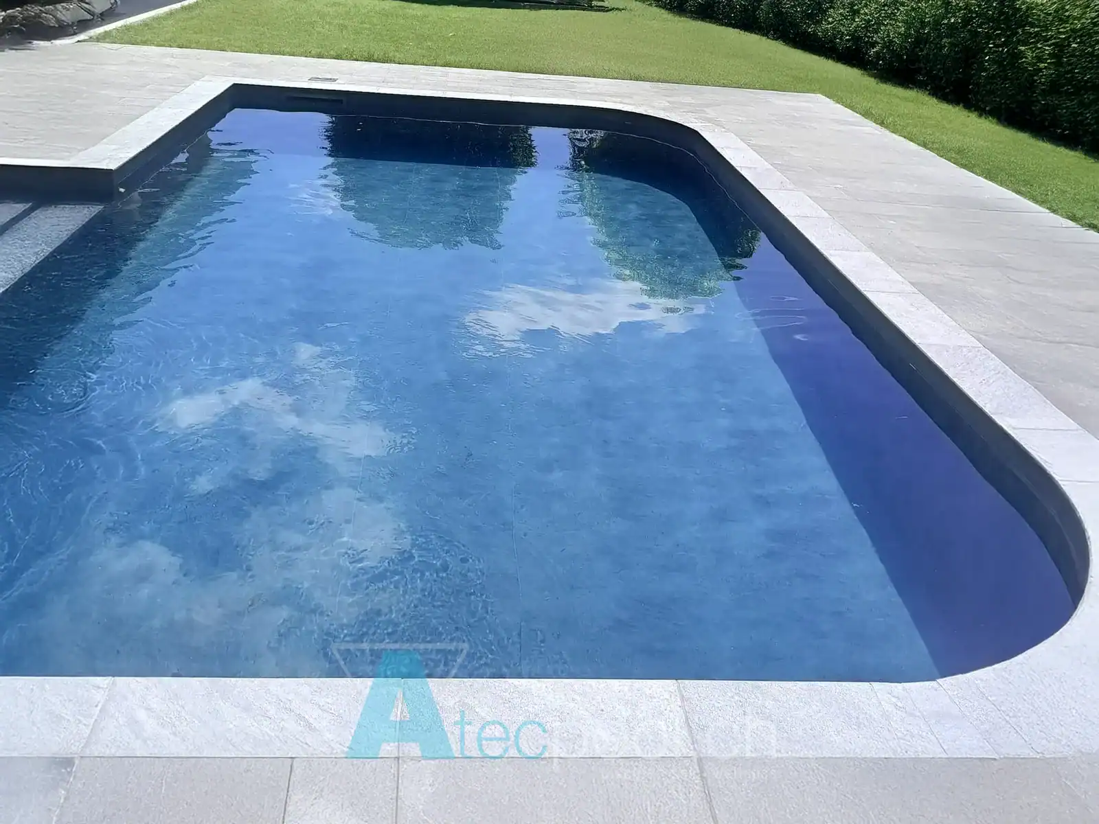 Piscina interrata sagomata con rivestimento blu e bordo in pietra – Atec Poollata Piscina interrata con forme arrotondate e rivestimento blu scuro in un giardino