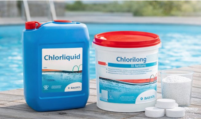 Cloro Depo di cloro liquido e pastiglie per il trattamento dell'acqua della piscina, posizionati su un bordo in legno vicino a una piscina all'aperto. Materiali plastici, colori blu, rosso e bianco, contenitori di grandi dimensioni.